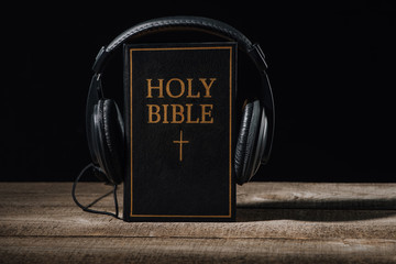 Wall Mural - Close-up shot of holy bible with headphones standing on wooden table isolated on black