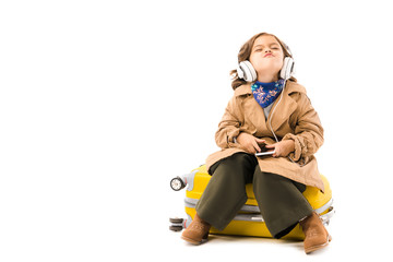 Wall Mural - grimacing little child in trench coat listening music with headphones and sitting on yellow suitcase isolated on white