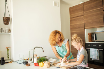 pretty little caucasian girl dressed in apron and wearing ponytail from blonde curly hair kneadling dough while cooking appetizing pie with mom, interior of modern wooden kitchen on background