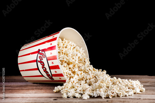 Fotografering Spilled box with popcorn on retro wooden desk and black background