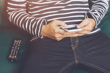 young man using television remote control and mobile smart phone is sitting Search for his favorite channel list on a sofa. on or off tv.	