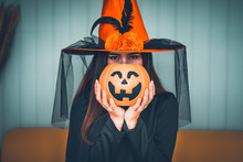 Attractive Beautiful Asian Woman Dressed As A Witch Holding Jack-o-lantern On Hand And Sitting On Yellow Sofa, Halloween Holidays In Cinematic Tone. 