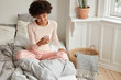 © WHstudio Leushin N - Joyful female blogger enjoys recreation time, does shopping online, uses mobile phone application, dressed in nightwear on comfortable bed, wears pyjamas, has Afro hairstyle. Leisure concept