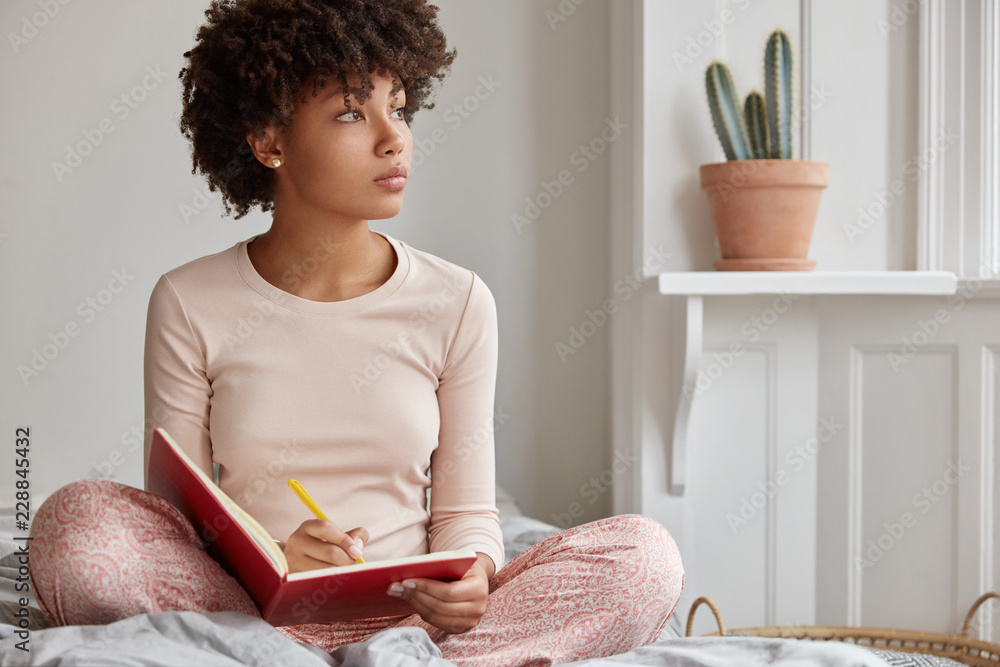 Cropped shot of dark skinned woman sits crossed legs, wears pyjamas, makes notes in diary, focused aside, blank copy space against domestic interior. Blogger creats publication for blog in notebook - obrazy, fototapety, plakaty 