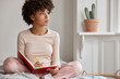 © WHstudio Leushin N - Cropped shot of dark skinned woman sits crossed legs, wears pyjamas, makes notes in diary, focused aside, blank copy space against domestic interior. Blogger creats publication for blog in notebook