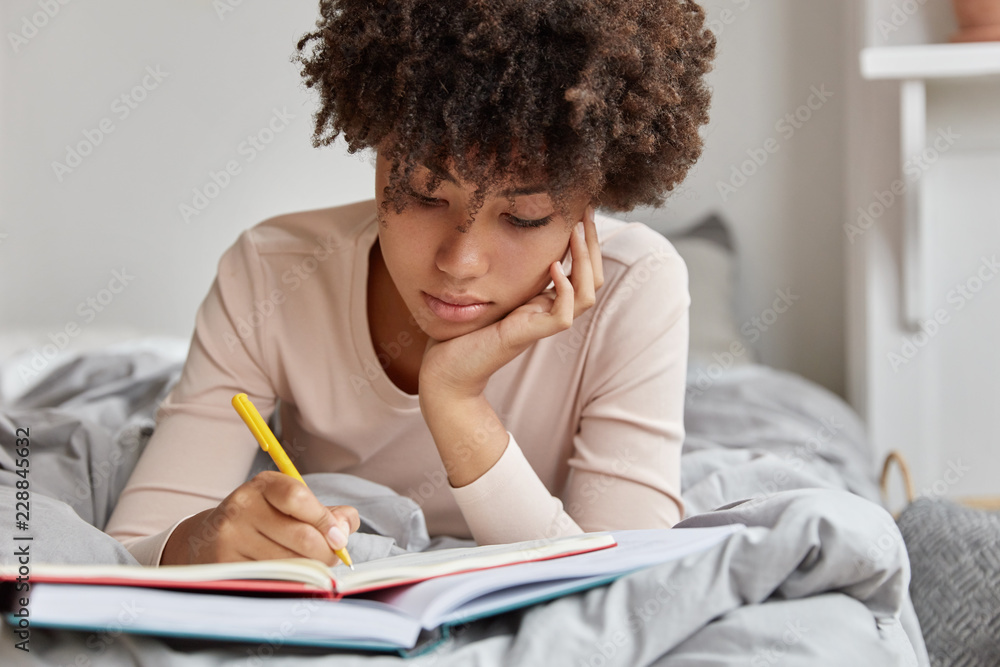 Image of beautiful woman with Afro hairstyle rewrites information in notebook from book, writes down homework, enjoys leisure time in bedroom, wears casual clothes. Female makes future plans - obrazy, fototapety, plakaty 