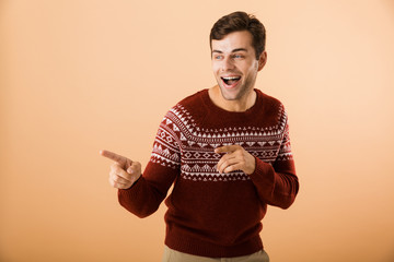 Canvas Print - Image of joyous man 20s with bristle wearing knitted sweater pointing finger aside at copyspace, isolated over beige background