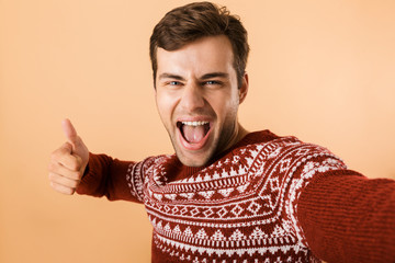 Canvas Print - Image of optimistic man 20s with bristle wearing knitted sweater laughing while taking selfie photo, isolated over beige background