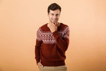 Canvas Print - Image of optimistic man 20s with bristle wearing knitted sweater posing on camera and touching chin, isolated over beige background