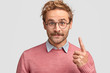 © WHstudio Leushin N - Horizontal shot of handsome satisfied hipster has mustache, dressed in elegant clothes, points with index finger upwards, wears round glasses, poses against white background. Man gives advice go there