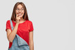 © WHstudio Leushin N - Smiling beautiful woman keeps index finger near mouth, looks myseriously aside, wears red casual t shirt and overalls, stands against white background with copy space for your promotional content