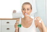 Fototapeta Łazienka - Woman brushing teeth and holding glass with mouthwash in bathroom