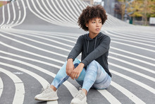 Outdoor Shot Of Pensive Dark Skinned Teenager Dressed In Casual Sportsclothes, Sneakers, Sits On Asphalt, Recreats After Having Sport Competitions, Has Fit Body. Teenagers And Active Lifestyle Concept