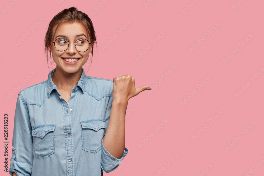 Attractive woman with happy expression advices use this copy space wisely, dressed in fashionable denim jacket, points with thumb aside, models against pink background. Go in this direction. - obrazy, fototapety, plakaty 