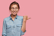 canvas print picture - Attractive woman with happy expression advices use this copy space wisely, dressed in fashionable denim jacket, points with thumb aside, models against pink background. Go in this direction.