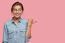 Attractive Woman With Happy Expression Advices Use This Copy Space Wisely, Dressed In Fashionable Denim Jacket, Points With Thumb Aside, Models Against Pink Background. Go In This Direction.