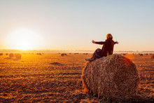 Happy Middle-aged Woman Sitting On Haystack In Autumn Field And Feeling Free With Arms Opened