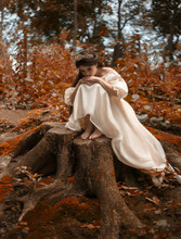 A Young, Sad Princess With Very Long Hair Sits On A Large Stump Of An Old Tree And Waits For Her Prince. The Girl Has A Vintage Dress And A Diadem. Artistic Processing, Unusual Colors.