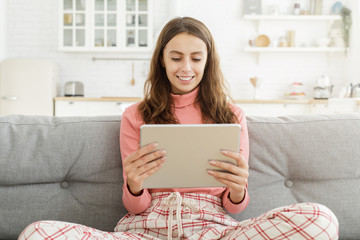 Young pretty smiling teen girl sitting on sofa, holding digital tablet computer with both hands, watching movie or using social media