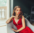 © kharchenkoirina - hot young adult, proud and imperious woman, dressed in a long scarlet red long dress, sexually holds her hand on her chest, sits on a grey chair in a loft room with window to the floor, cheeky style