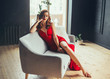 © kharchenkoirina - hot young woman, dressed in a long scarlet red long dress, sexually shows her bare legs sitting on a grey sofa in a loft modern room with window to the floor. Jessica rabbit halloween image