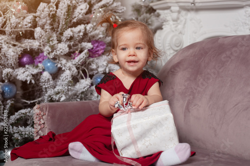 A Beautiful Cute Little Girl Dressed In An Elegant Evening Red Dress Sits On The Couch And Opens A New Year S Gift With Christmas Tree On The Background Stock Photo Adobe