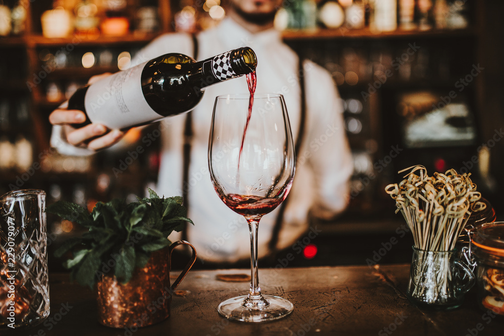 Close up shot of a bartender pouring red wine into a glass. Hospitality, beverage and wine concept. - obrazy, fototapety, plakaty 