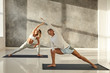 Young couple practicing yoga together. Indoor picture of handsome tanned guy on mat doing standing pose s to strengthen legs, stretching arms and looking up, blonde woman doing the same in background