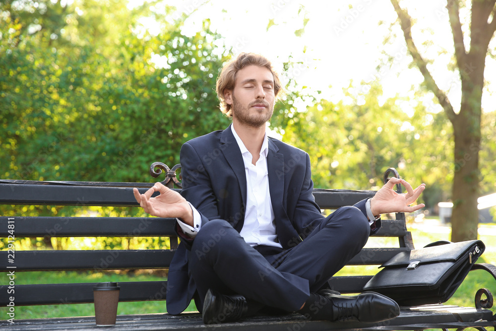 Handsome young businessman meditating on bench in park