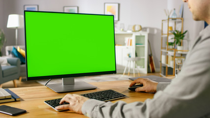 Over the Shoulder Shot of Professional Freelancer Working on Green Mockup Screen Personal Computer From Home. Man's Hands Typing, He's Browsing Through the Internet, Using Computer In His Living Room.