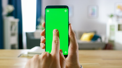 Wall Mural - Close-up of a Man Holding Green Mock-up Screen Smartphone and Using Touchscreen Gestures. Touching Mobile Phone Chroma Key Screen. In the Background Cozy Homely Atmosphere.