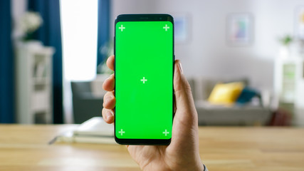 Wall Mural - Close-up of a Man Holding Green Mock-up Screen Smartphone. Mobile Phone has Chroma Key Screen. In the Background Cozy Homely Atmosphere.