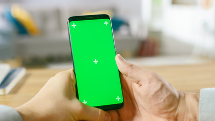 Wall Mural - Close-up of a Man Holding Green Mock-up Screen Smartphone in His Hands. Using Wireless Device to Browse Through Internet. In the Background Cozy Living Room or Home Office.