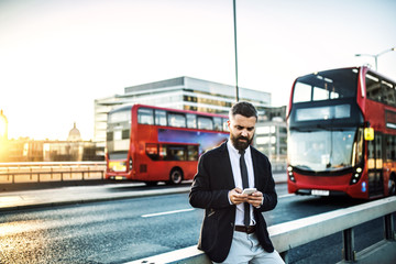 Wall Mural - Hipster businessman standing by the road in London, using smartphone.