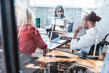 Wall Mural - view through glass of young businessman in wheelchair working with colleagues in office