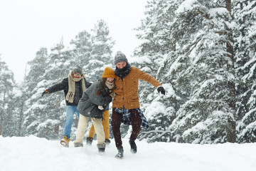 Positive excited young friends in warm clothing running over winter hill while having fun in forest