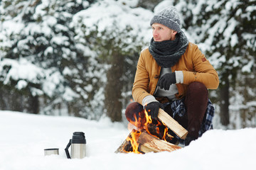 Pensive handsome young male hiker in warm scarf sitting by campfire while lighting it and adding timbers in winter forest, he looking into distance