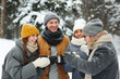 © pressmaster - Happy excited young travelers in winterwear standing in forest and drinking hot tea while chatting at winter hike