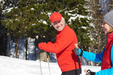 Wall Mural - Happy senior sportsman looking at his spouse while both moving on skis or trekking in winter forest