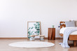 Floral graphic in frame next to wooden bedside table and bed with pillows and blankets, white round carpet on the floor, copy space on the wall