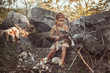 Caveman, manly boy making primitive stone weapon. Funny young primitive boy outdoors near bonfire. Evolution survival concept. Calm boy outside sitting at his rocky settlement. Prehistoric tribal man