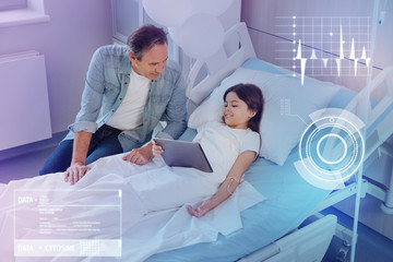 New technologies. Emotional little girl recovering and holding a modern tablet while staying in a hospital bed and talking to her kind curious father