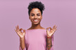 © antianti - Studio shot of pleased young beautiful African American female shows ok sign, dressed in striped casual jacket, being in good mood, poses against lavender background. People and approval concept