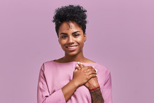 Portrait Of Kind Hearted African American Female In Fashionable Overalls, Keeps Hands On Chest, Shows Her Kindness And Sympathy, Has Pleased Cheerful Expression, Isolated Over Lavender Background