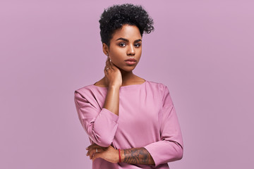 Gorgeous dark skinned young female with Afro hairstyle and confident look, poses for fashionable magazine, looks aside thoughtfully, wears trendy shades, poses against lavender studio background