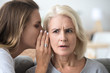 Young woman whisper in aged mother ear sharing secret or hidden information, surprised elderly mom hearing shocking news from adult daughter, parent and kid being best friends gossiping at home