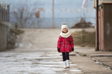 Fool-length portrait of cute little young funny pretty child girl in nice warm winter clothing walking confidently alone on white bright blurred outdoors copy space background. Outdoors activities.