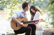 © soleg - young couple sitting on a log by the river and playing guitar, summer nature, bright sunlight, shadows and green leaves, romantic feelings