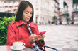 © Maridav - Cafe woman using mobile phone drinking cappuccino coffee at outdoor street european city. Europe travel lifestyle. Asian businesswoman in fashion red trench coat.