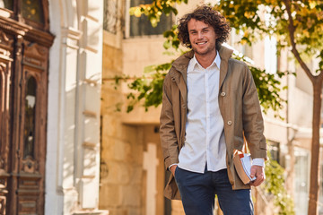 Wall Mural - Outdoor image of handsome young man with books outdoors. College male student carrying books in college campus in autumn street background. Smiling cheerful guy with curly hair posing with books.